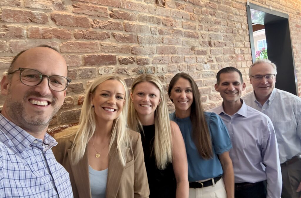Group selfie of six people smiling in front of an exposed brick wall
